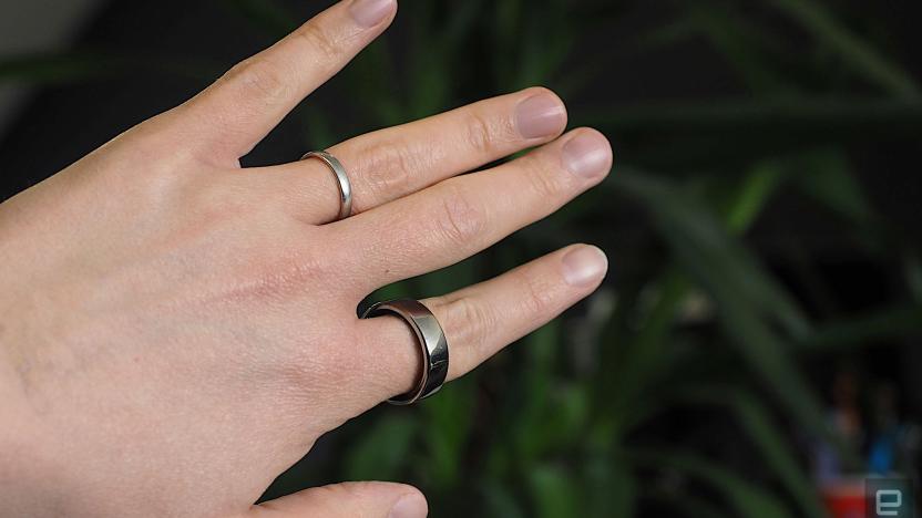 Image of a hand wearing an Oura Ring on the index finder in front of a dark green plant.